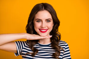 young woman smiling against an orange background, hand under her chin propping up her perfect white smile
